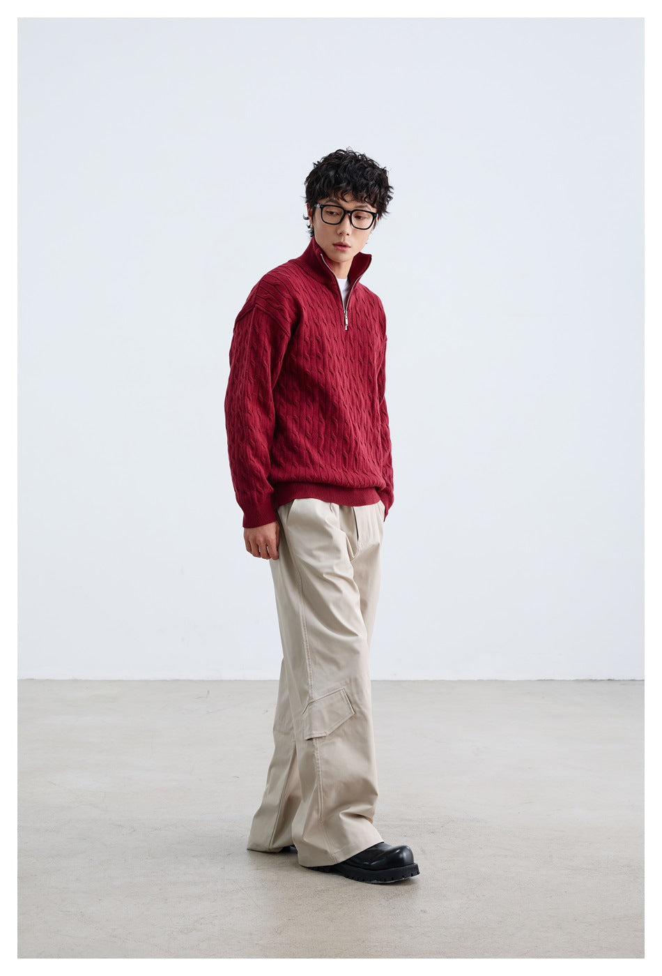 Model wearing a burgundy cable-knit quarter-zip sweater with a raised collar and loose beige trousers.