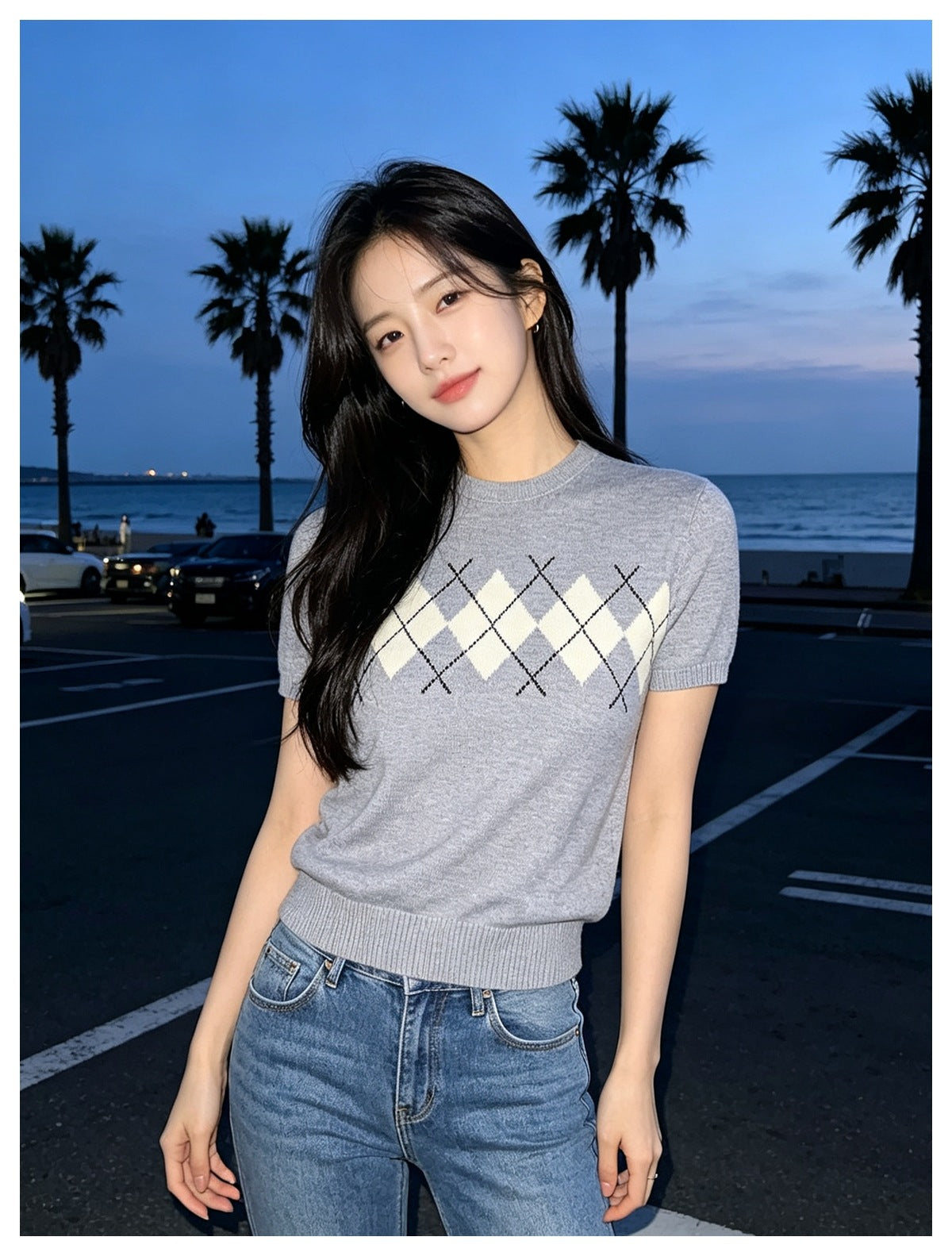 Woman wearing a heather gray short-sleeve argyle knit top with blue jeans, standing by the beach at dusk