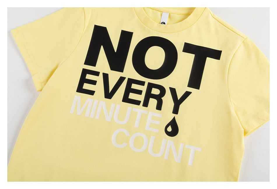 Flat-lay front of a pale yellow short-sleeve T-shirt with bold black and white “NOT EVERY MINUTE COUNT” lettering and a