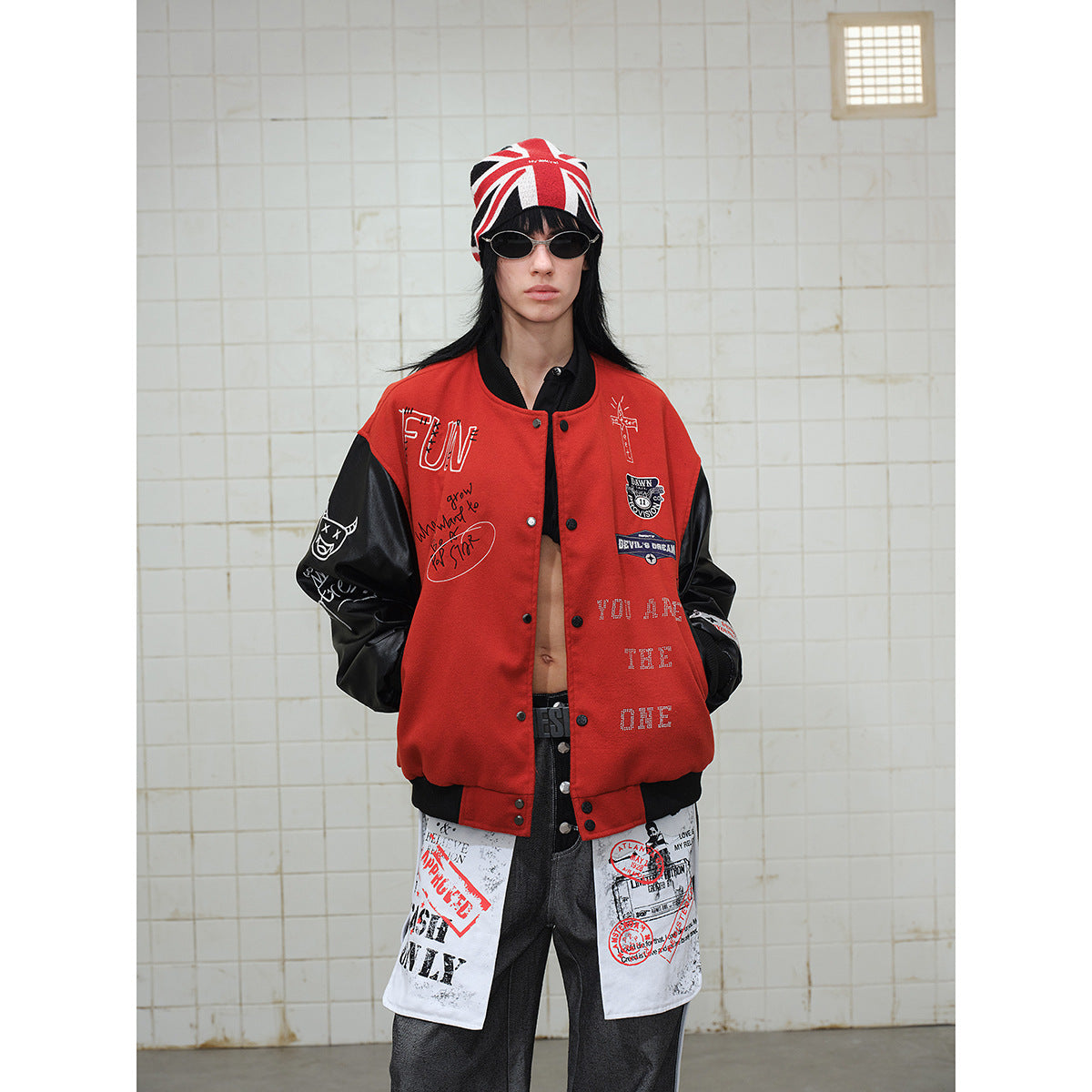 Model wearing a red varsity jacket with black sleeves and graphic patches, paired with gray trousers and a Union Jack beanie.