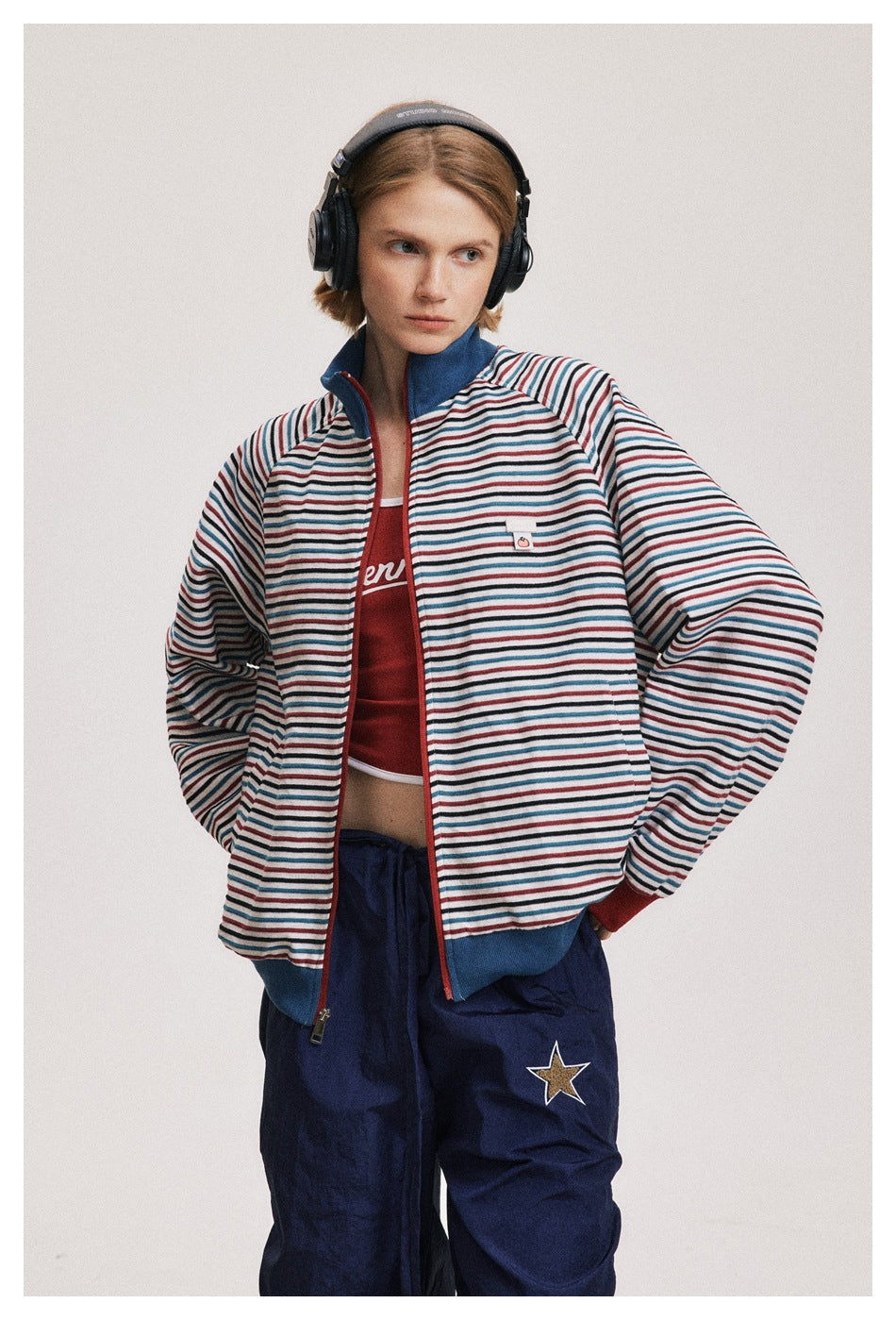 Model wearing an open striped zip jacket with blue trim and red cuffs, showing a red cropped top underneath.