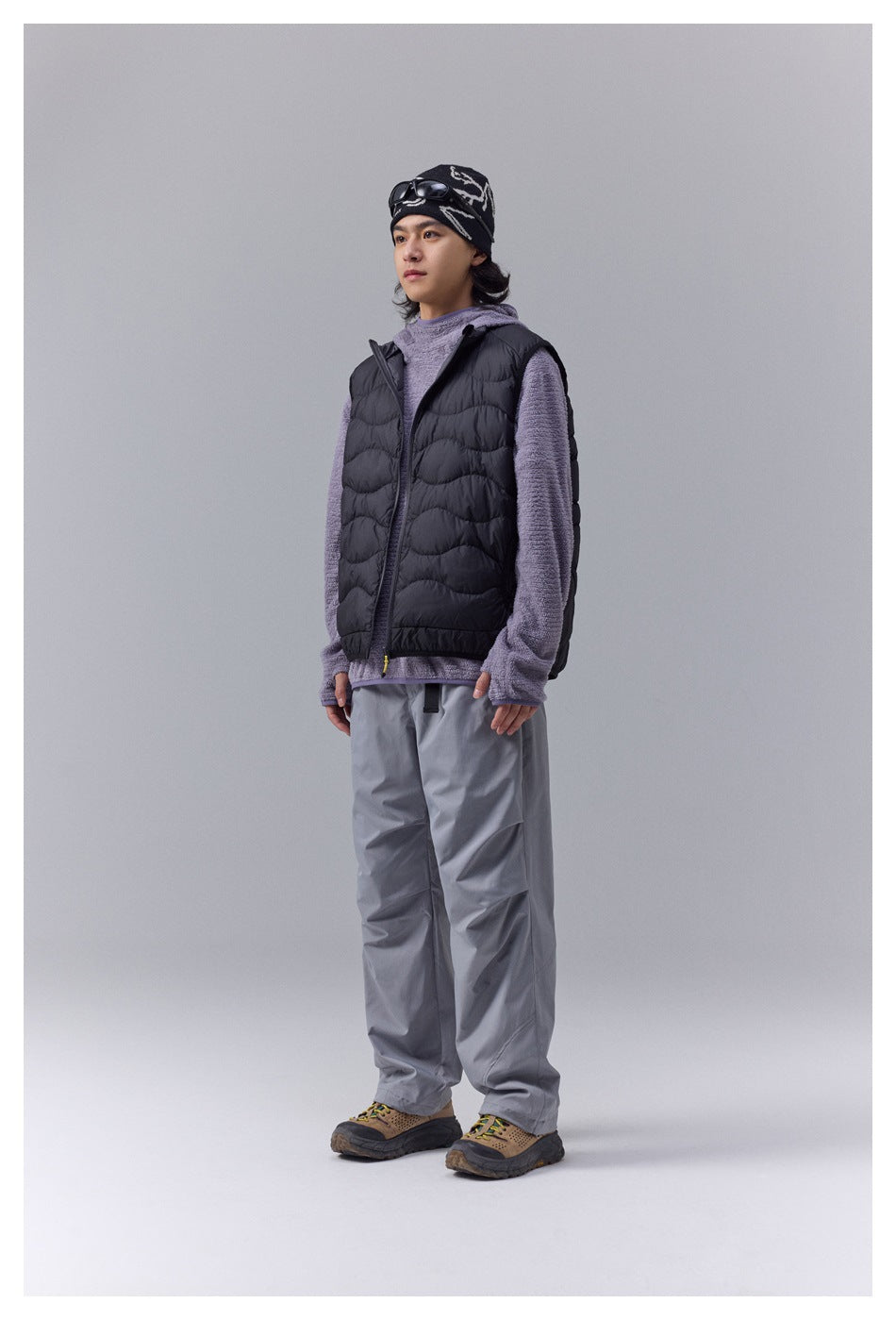 Model wearing a black quilted zip vest over a purple fleece-like top with gray pants and a patterned beanie