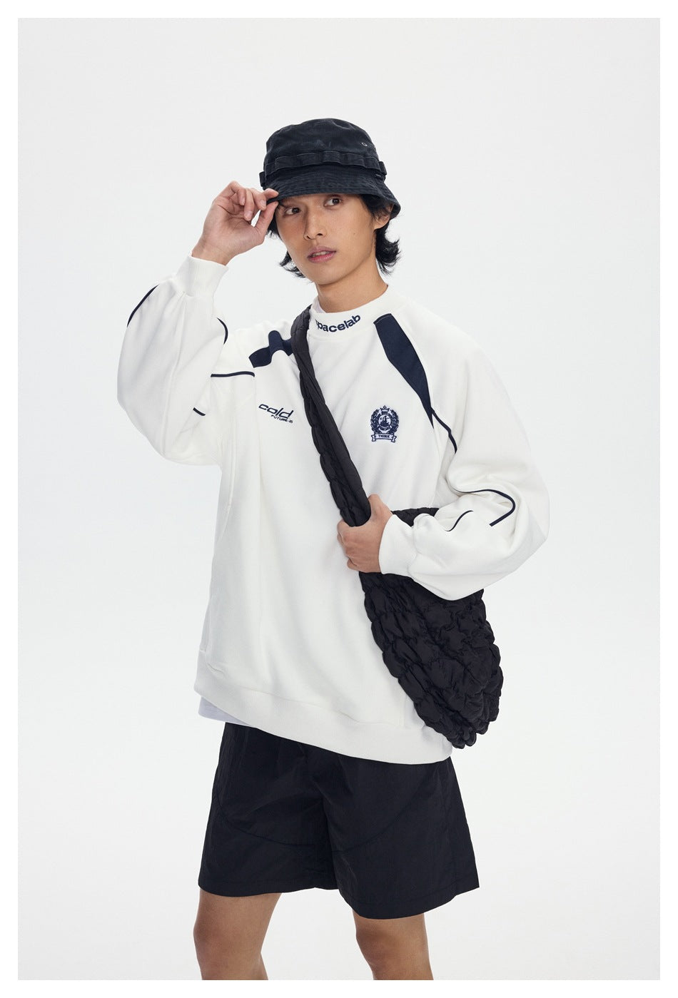 Model wearing a white sweatshirt with navy accents and chest graphics, black shorts and a black crossbody bag.