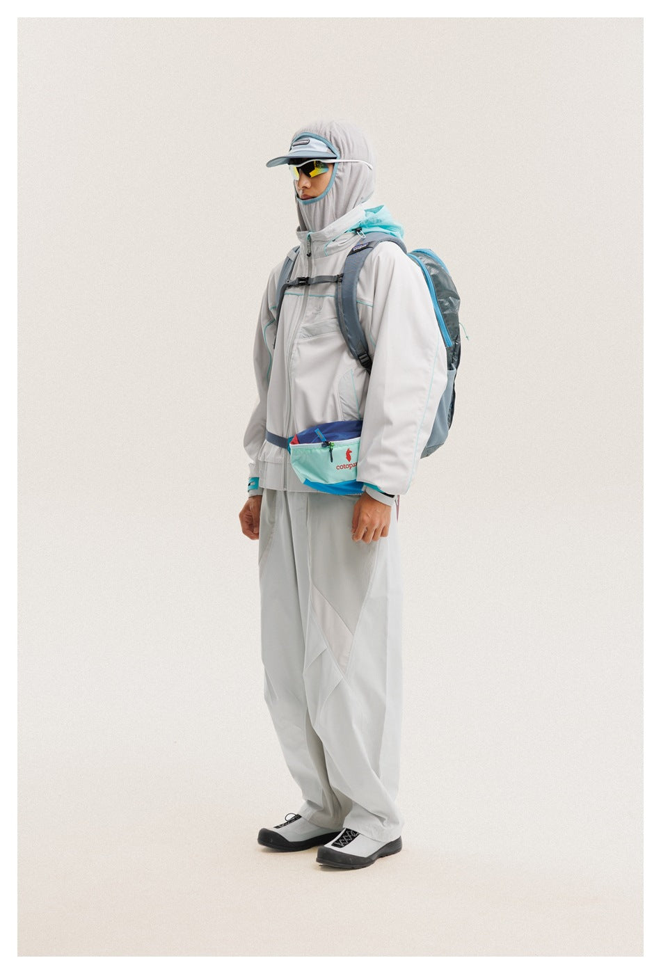 Three-quarter side view of a model wearing a light gray outdoor jacket and pants with turquoise accents, cap, hood and