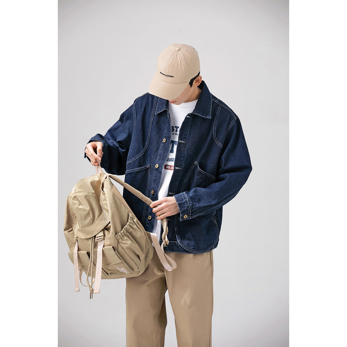 Model wearing a dark indigo denim jacket and beige trousers while holding a beige backpack.
