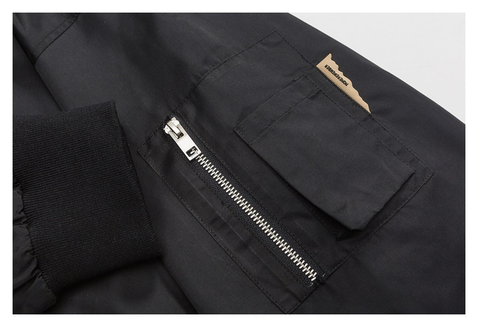 Close-up of a black garment showing a silver zipper, flap pocket, ribbed cuff, and small tan label.