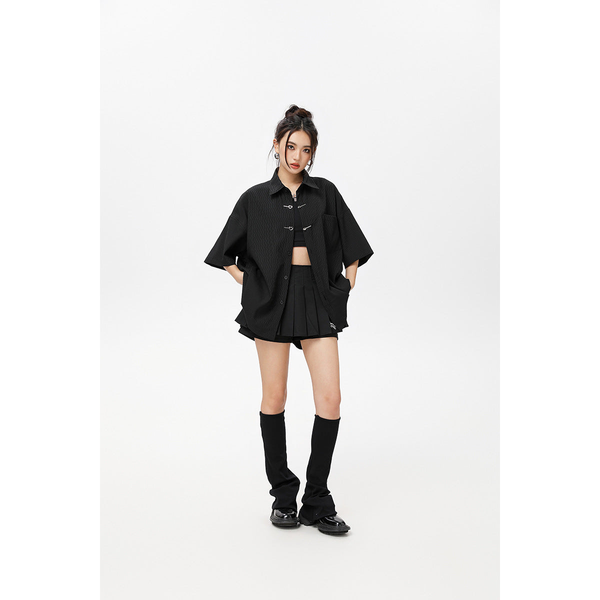 Model wearing an oversized black short-sleeve shirt with metal toggles and a black pleated skirt.