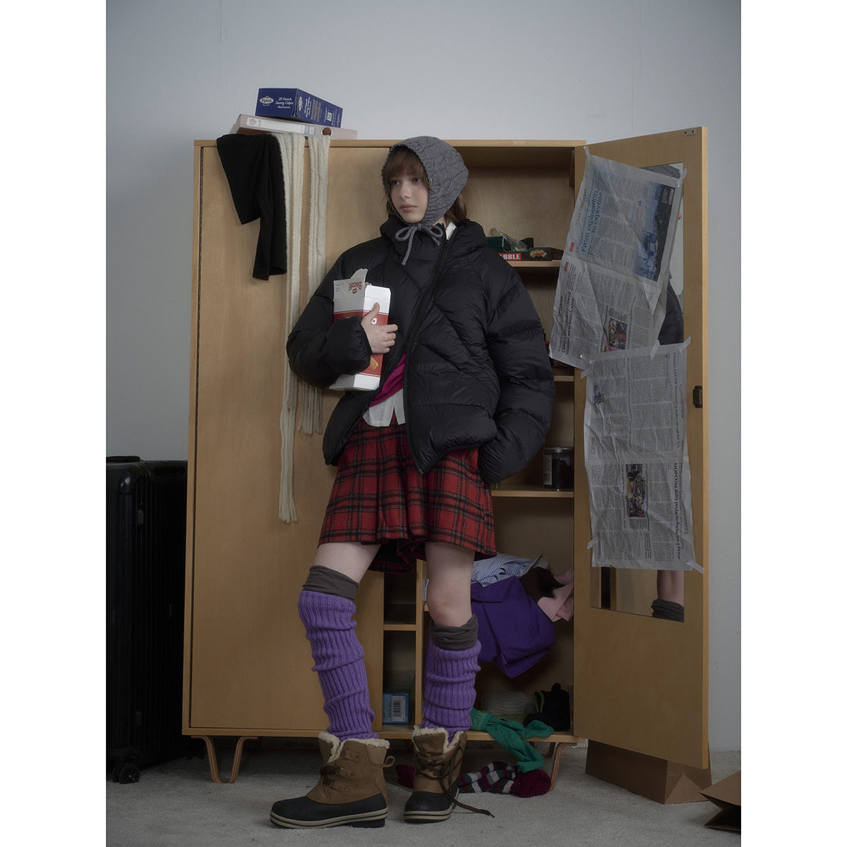 Full-length view of a model in a black puffer jacket, gray bonnet, red plaid skirt, and purple leg warmers.