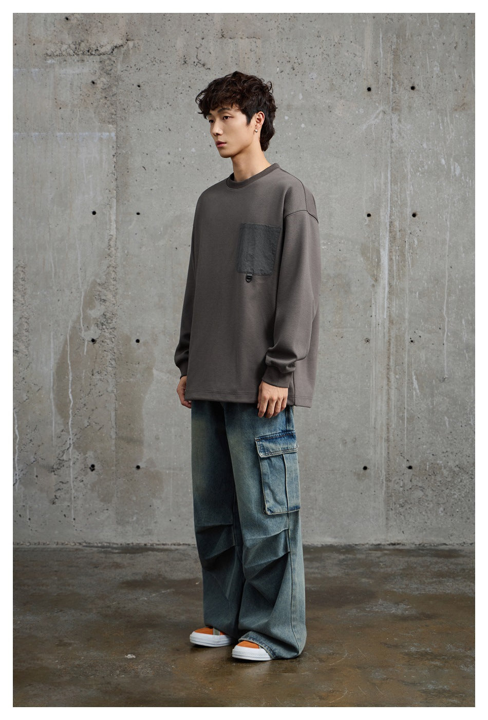 Model wearing an oversized charcoal-gray long-sleeve top with a dark chest pocket and loose blue cargo jeans, front view.