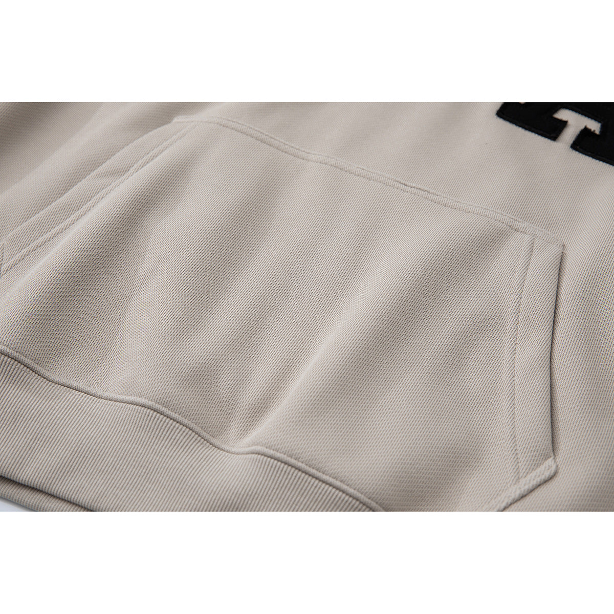 Close-up of the kangaroo pocket and fabric on a light beige hoodie, with part of the black chest lettering visible.