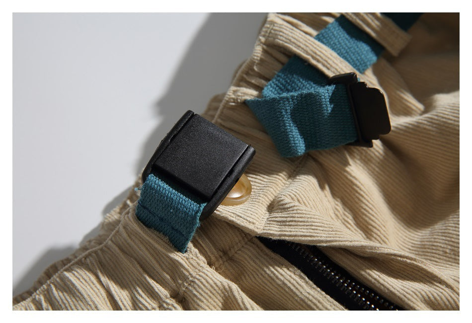 Close-up of teal webbing tabs and black buckles beside the zipper on tan corduroy pants.