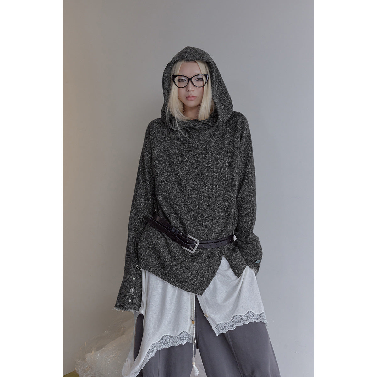 A standing model wears a charcoal hooded top with an angled black belt, layered white lace-trim shirt and loose gray pants.