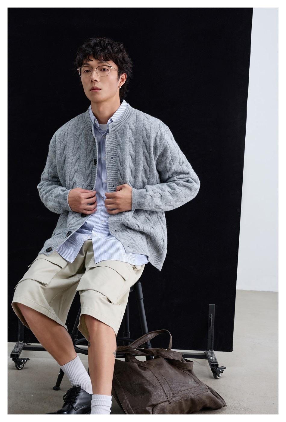 Seated person wearing a light gray cable-knit button cardigan over a striped shirt and beige shorts.