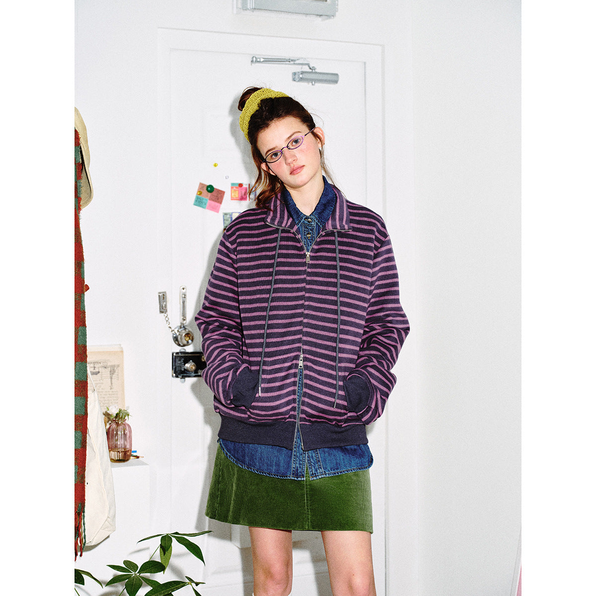 Model wearing a purple and black striped zip-front jacket with a denim shirt and green skirt, shown from the front.