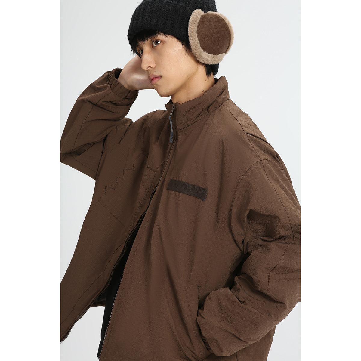 Model wearing a brown zip-front jacket with a high collar and elastic cuffs, paired with a black knit earflap hat.