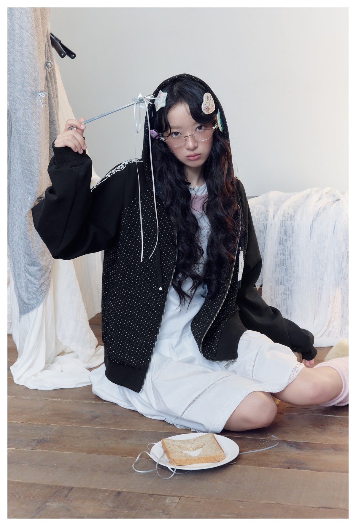 Seated model wearing an open black dotted hooded jacket with white sleeve graphics over a white dress.
