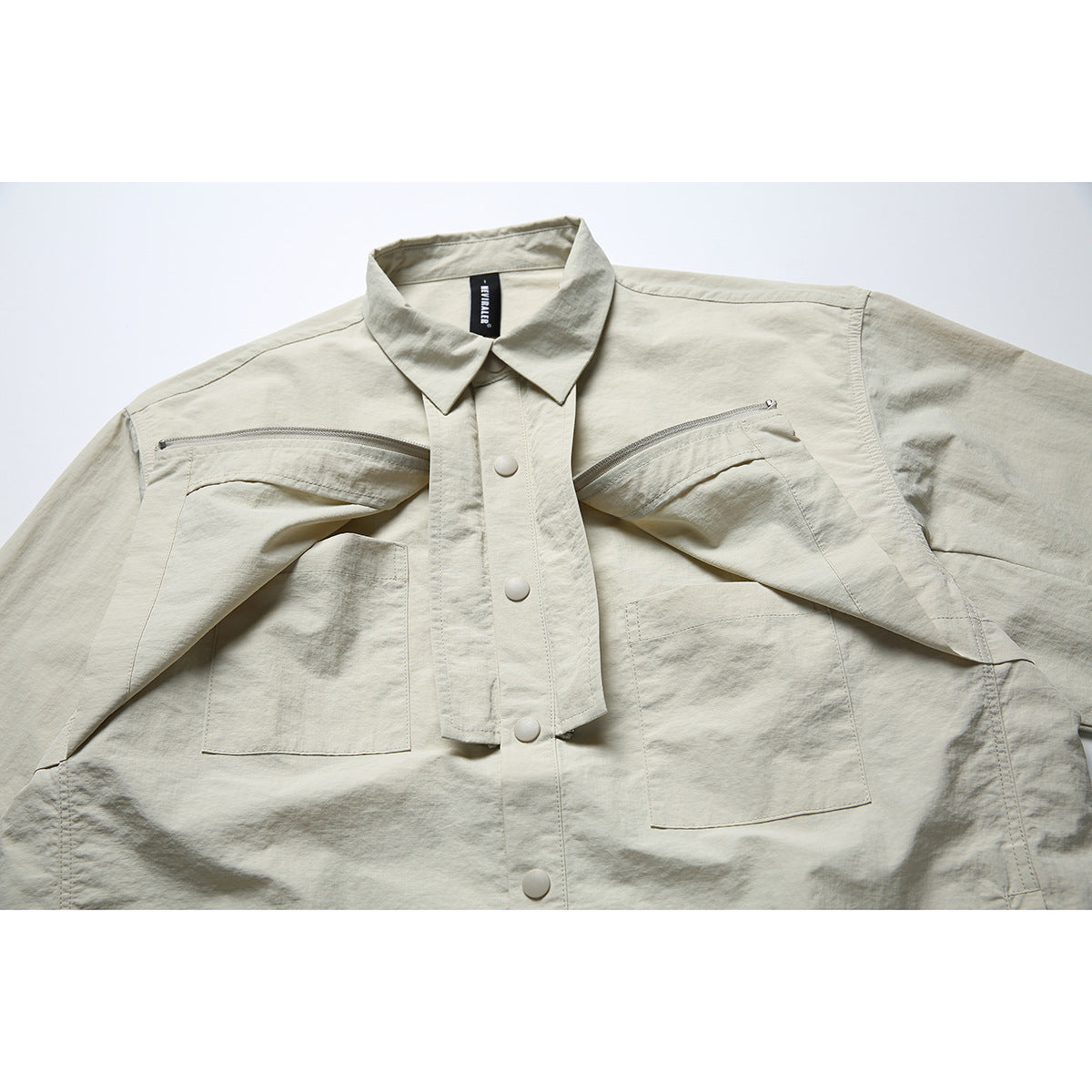 Close-up of a pale beige shirt jacket with a pointed collar, snap-button placket, chest pockets and curved zip openings.