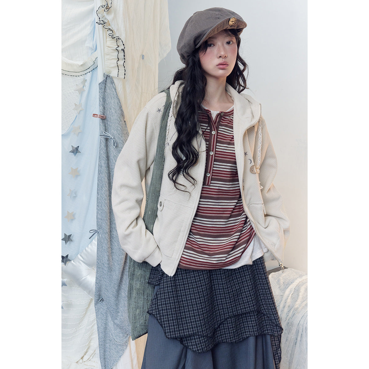 Model wearing an ivory zip hoodie with celestial motifs over a burgundy striped top and layered dark skirt.