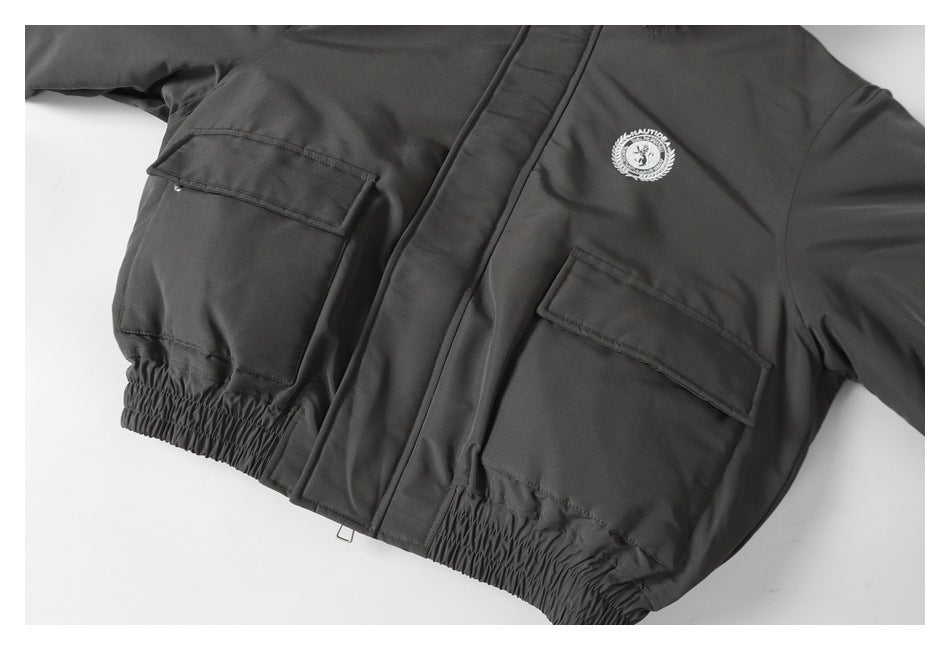 Close-up of a charcoal jacket front with flap pockets, an embroidered chest emblem, and an elasticated hem.