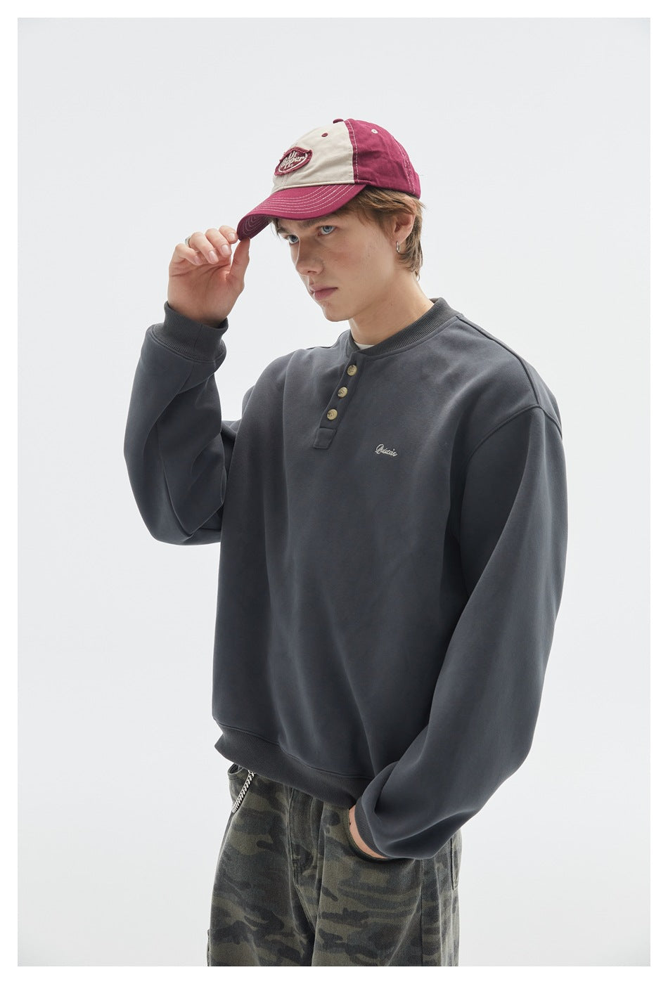 Close view of a model wearing a charcoal three-button sweatshirt with small chest embroidery and a burgundy-and-cream cap.