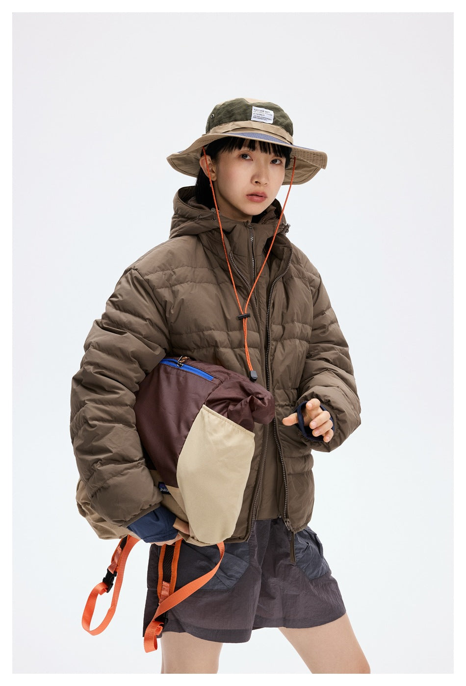 Model wearing a brown hooded puffer jacket and gray shorts, holding a two-tone tote bag.