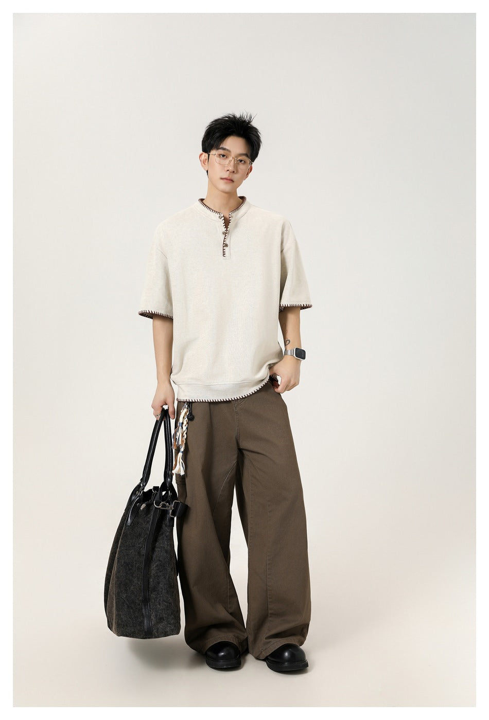 Full-length model wearing an ivory contrast-stitched henley, wide brown trousers, and carrying a black tote.