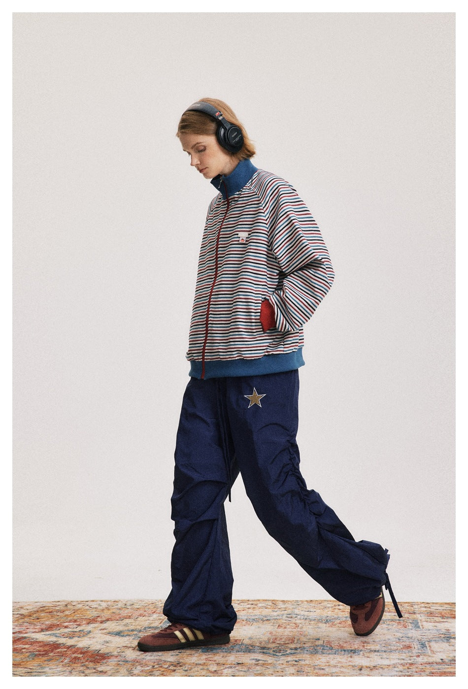 Model wearing a striped zip jacket with blue stand collar and ribbed hem, navy parachute pants and headphones, walking