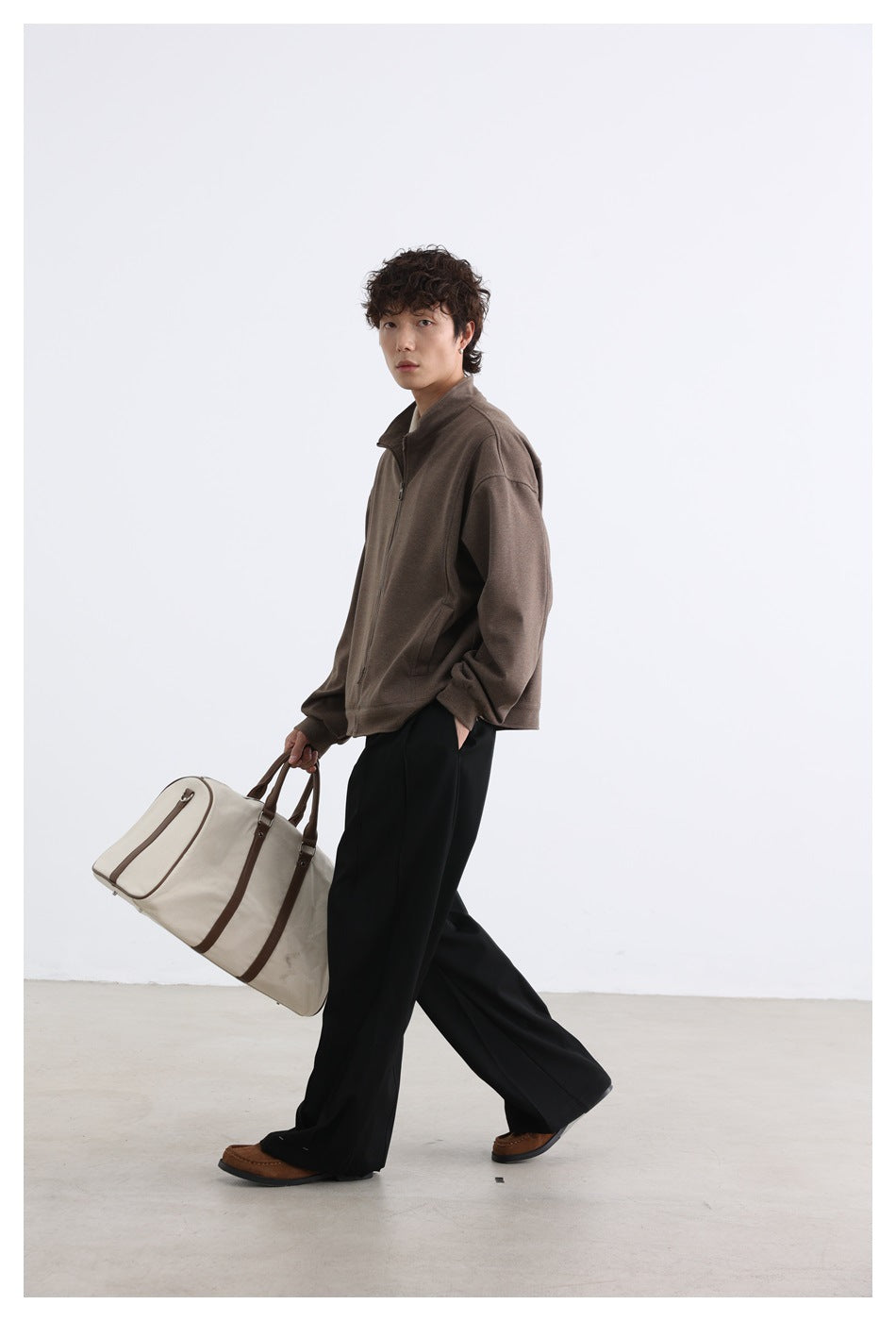 Model walking in a brown zip-front jacket and carrying a large cream duffel bag.