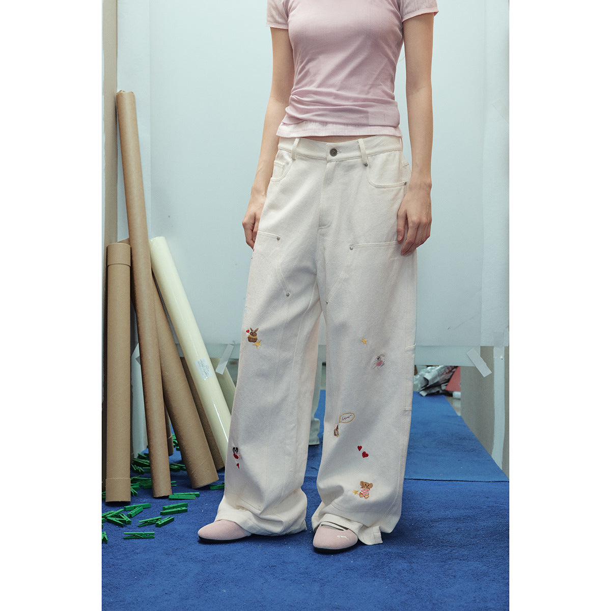 Front view of a model wearing ivory wide-leg pants with utility pockets and scattered colorful embroidered motifs.