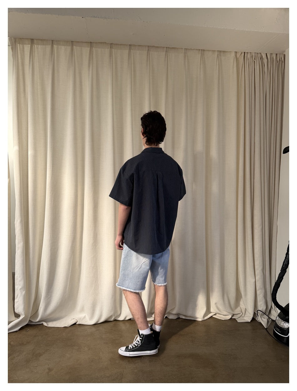 Back view of a model wearing a dark pinstriped short-sleeve shirt and light blue frayed denim shorts, framed by a white