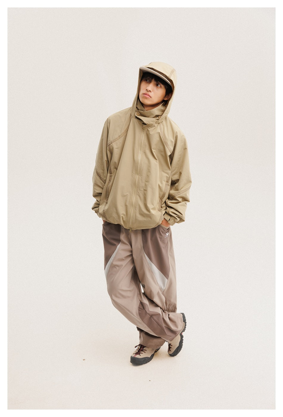 Model wearing a tan hooded zip jacket and taupe color-block pants, with the hood up.