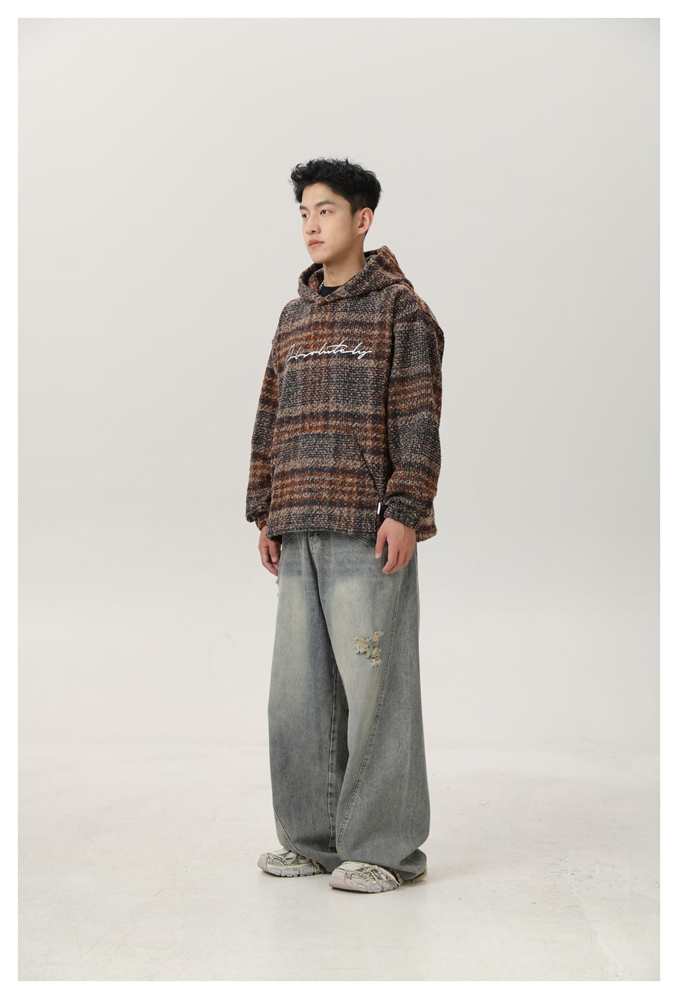 Man wearing plaid hooded top with front pouch pocket and wide-leg faded jeans, angled side view