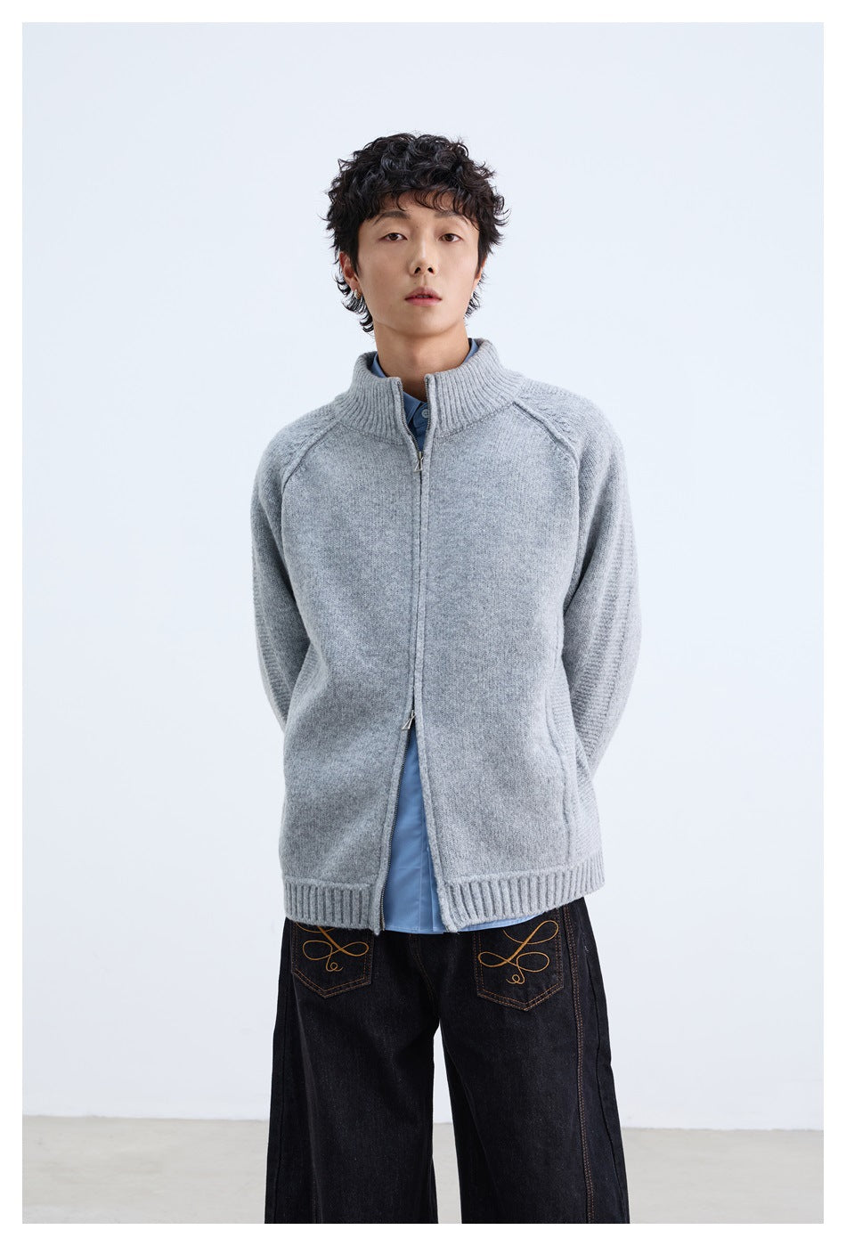 Model wearing a light gray zip-front knit cardigan with a ribbed stand collar over a blue shirt and black jeans.