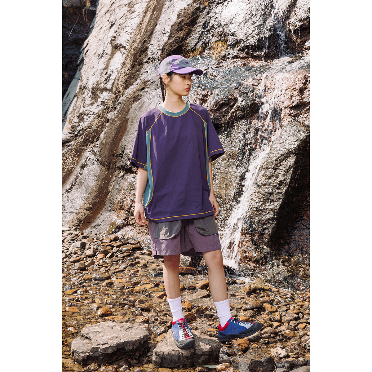 Model wearing a purple short-sleeve shirt with teal panels and yellow piping, paired with purple shorts outdoors by rocks and