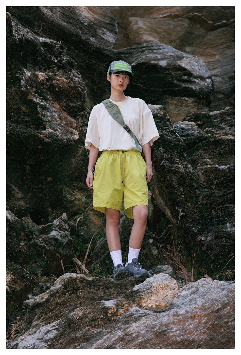 Model in a cream T-shirt with chest pocket, yellow shorts, cap and crossbody bag, standing on rocks