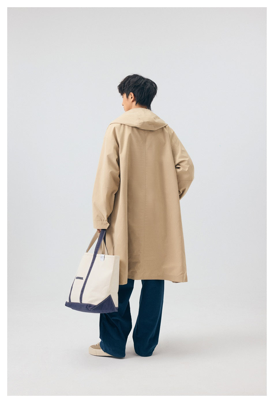 Rear view of a model wearing a long beige hooded coat with navy trousers and carrying a cream tote.