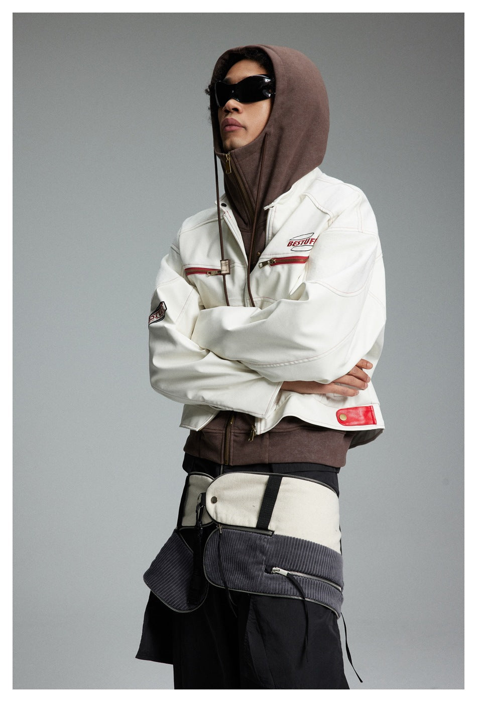 Model in a white red-trimmed jacket over a brown hooded layer, arms crossed above black trousers.
