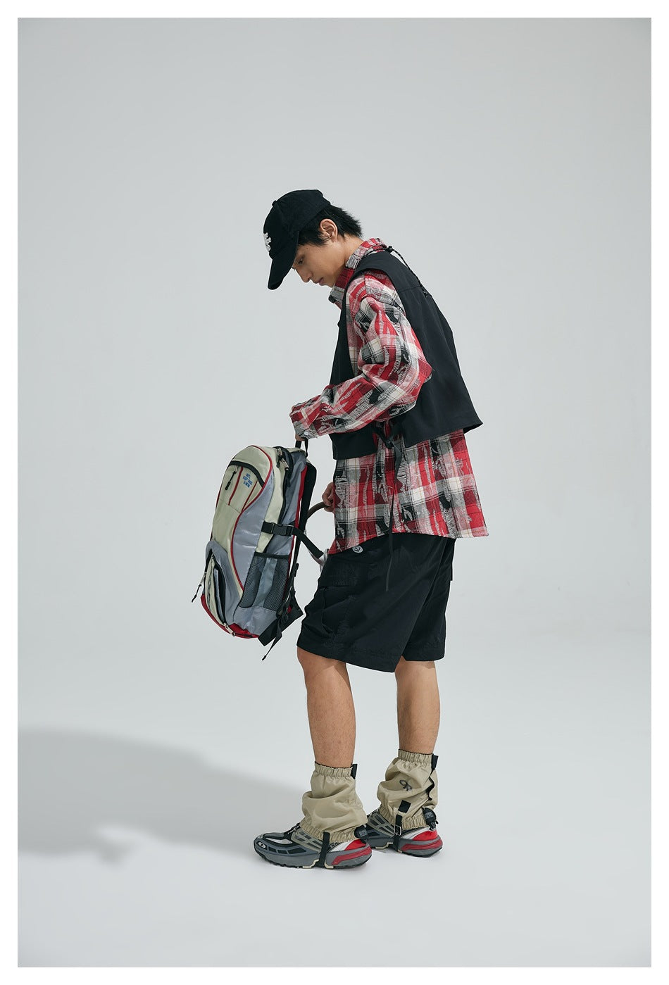 Side view of a model wearing a black utility vest over a red plaid shirt with black shorts, holding a backpack