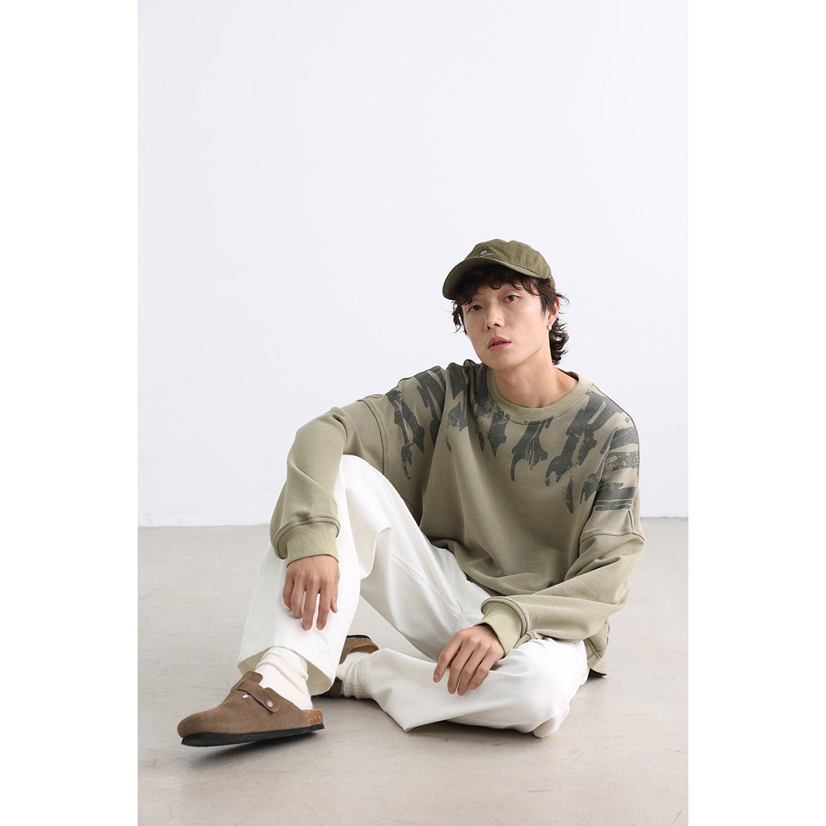 Seated model wearing a khaki sweatshirt with dark abstract shoulder graphics and white pants.