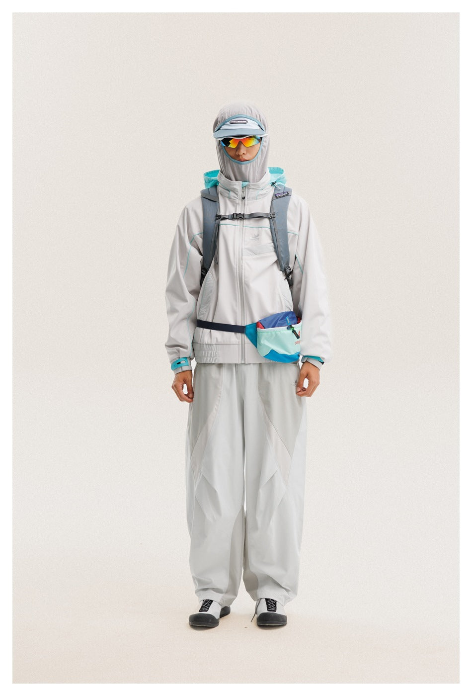 Front view of a model in a light gray jacket and loose pants with turquoise trim, hood, cap, sunglasses and crossbody pouch.