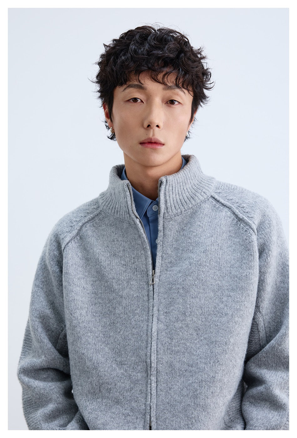 Close view of a model wearing a light gray zip-front knit jacket with a ribbed stand collar over a blue shirt.