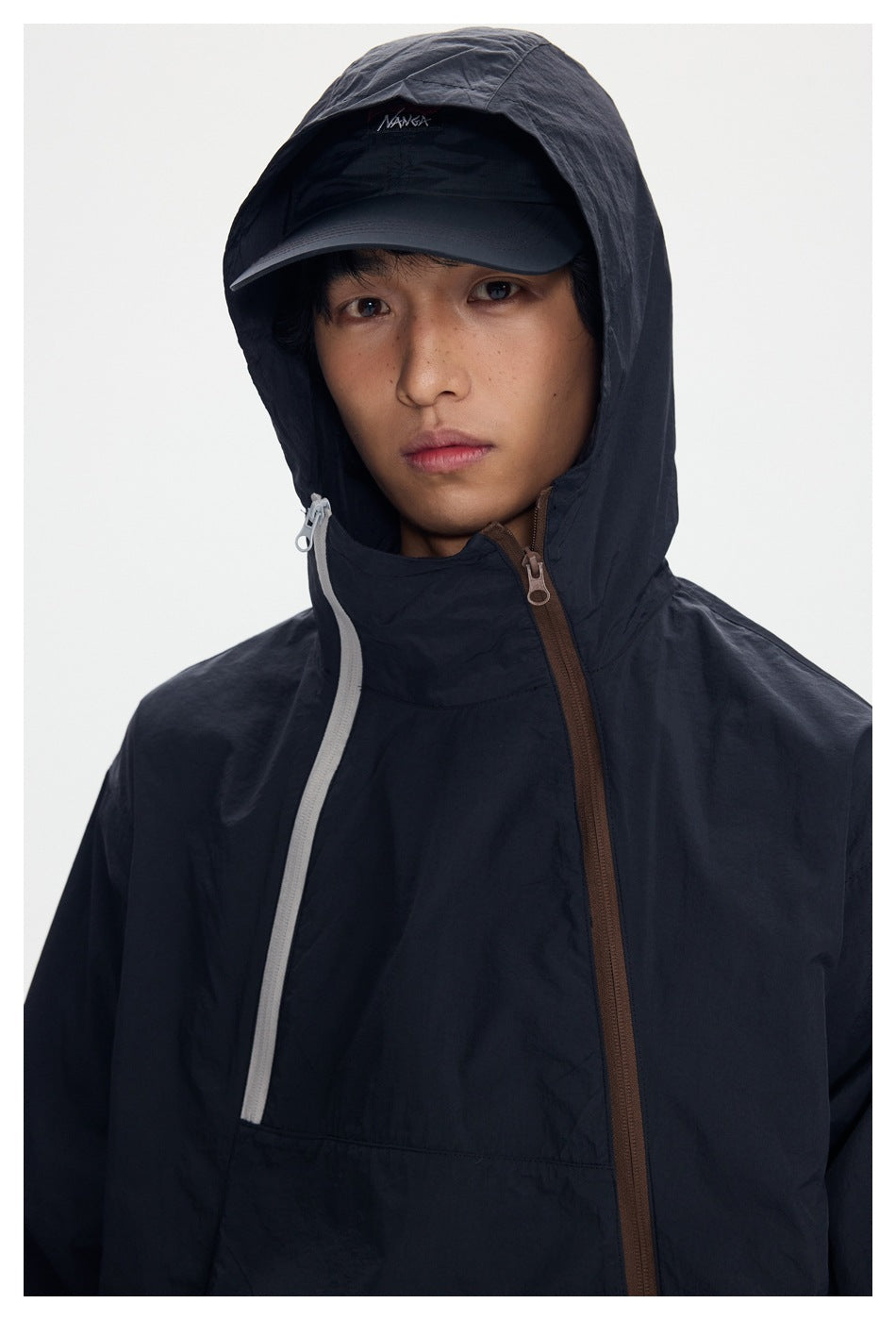 Close-up of a model wearing a dark hooded jacket with a brown front zipper and a curved light-colored chest zipper.