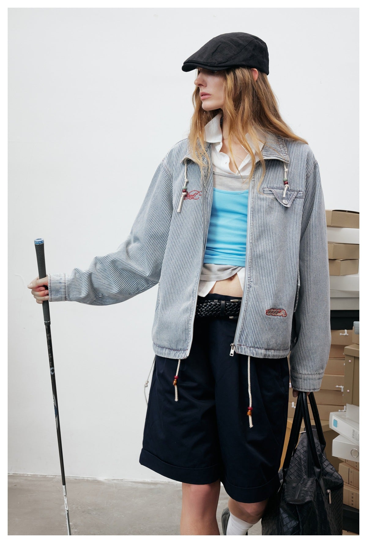 Close three-quarter view of a model in the striped zip jacket with beaded drawcords, navy shorts, cap and black woven bag