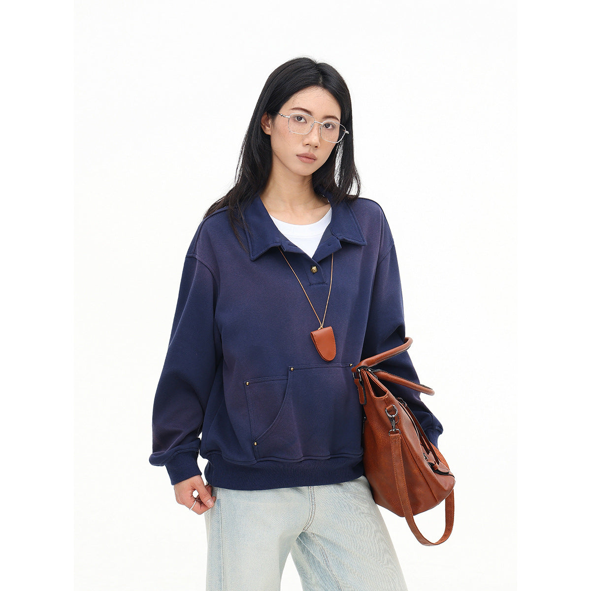 Closer view of a model in an oversized navy collared pullover with a pouch pocket, pale jeans, and a brown handbag.