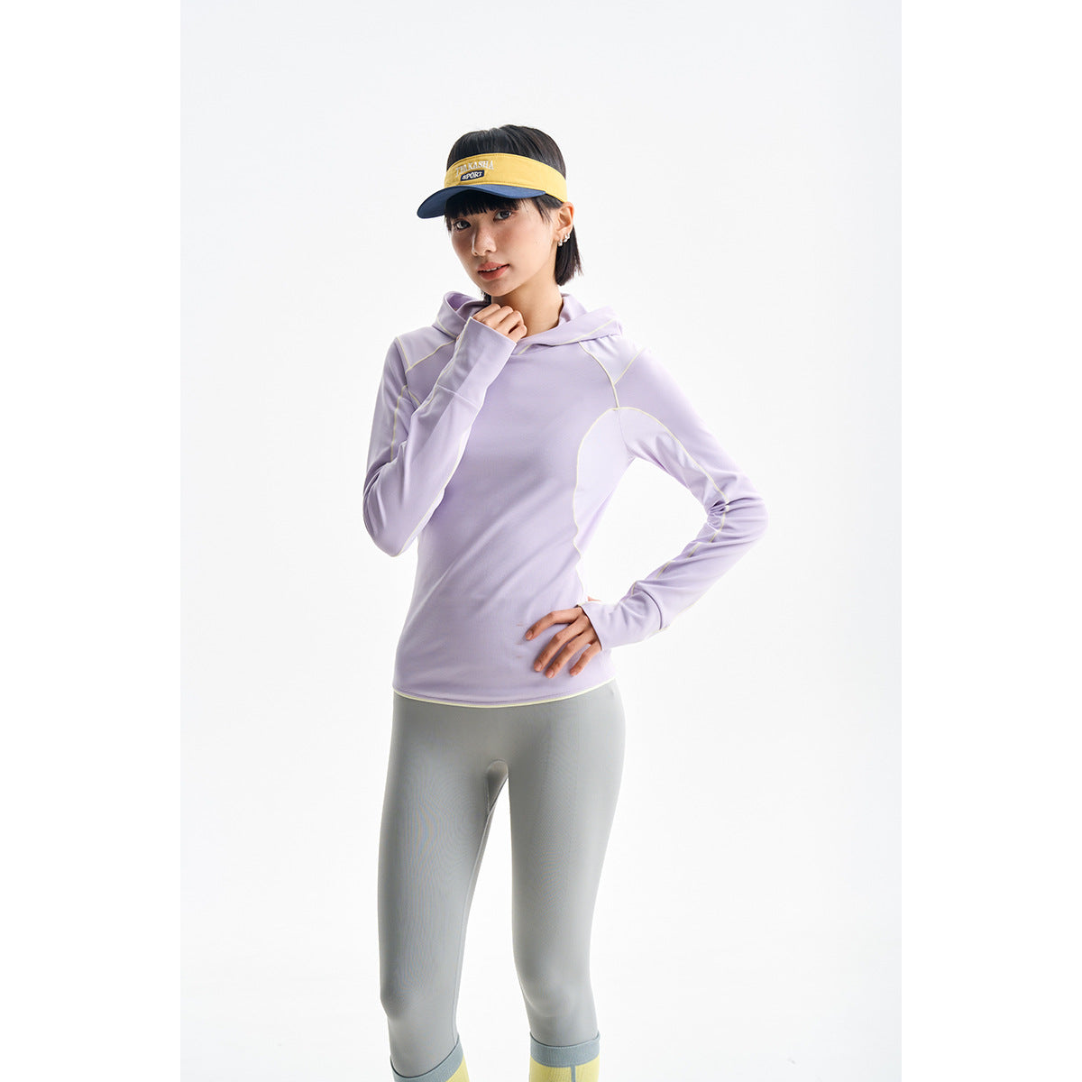 Model wearing a lavender hooded athletic top with pale contrast piping, gray leggings, and a yellow visor.