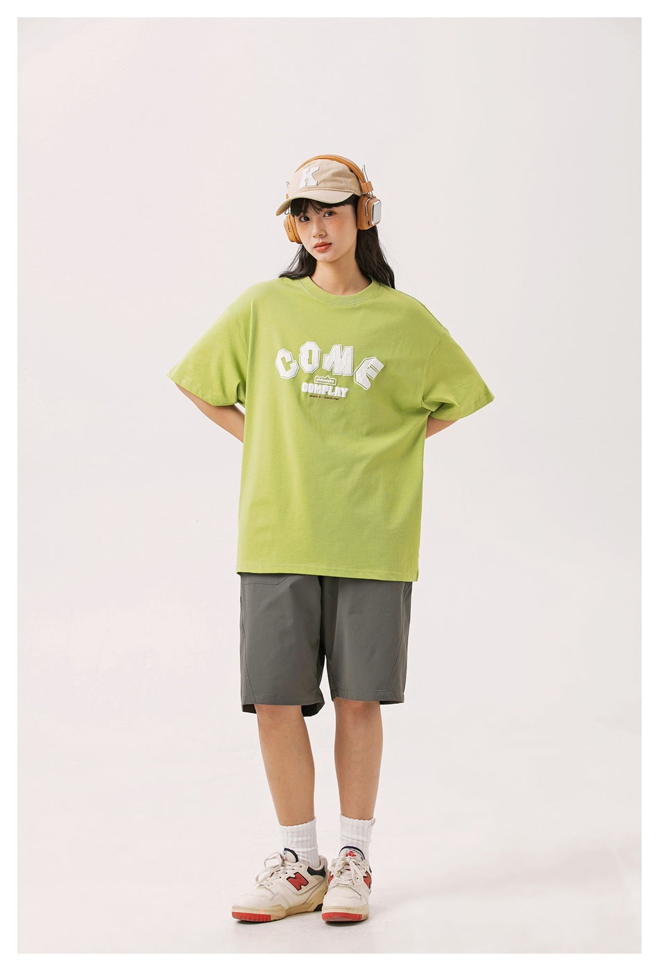 Model wearing a lime green oversized graphic T-shirt with gray shorts, beige cap, and headphones, shown full length from the