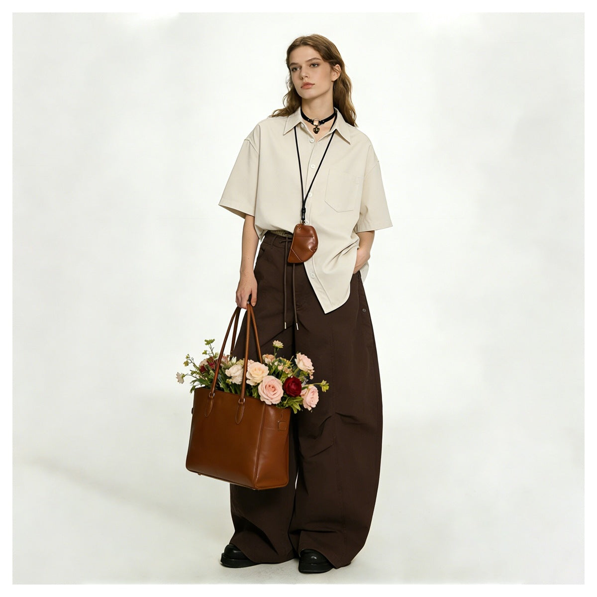 Model wearing brown wide-leg pants with a beige short-sleeve shirt, holding a tan tote bag with flowers