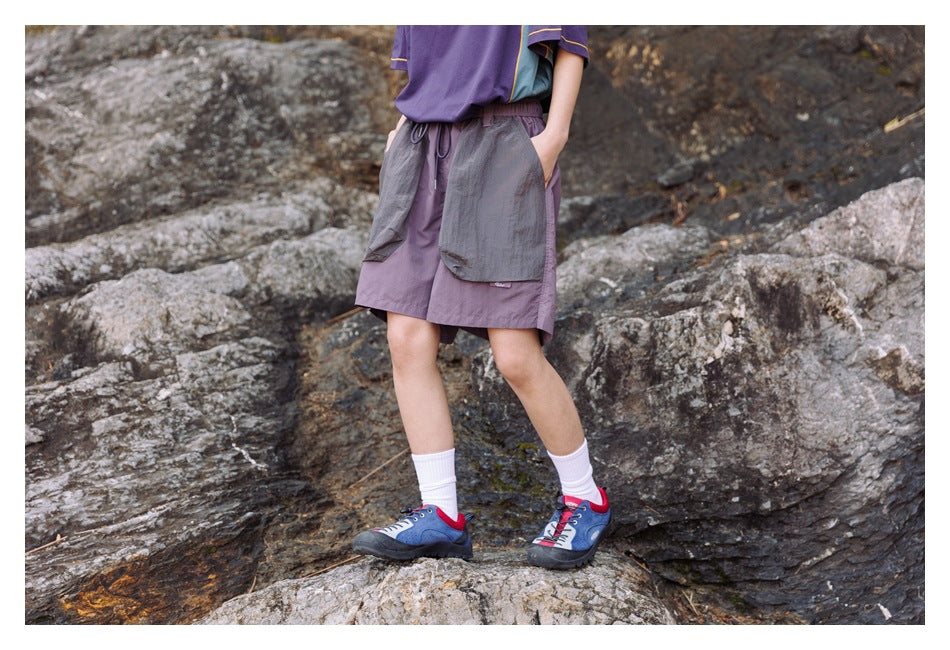 Person in purple shorts with large gray patch pockets and a hand in the pocket, standing on rocks, cropped view
