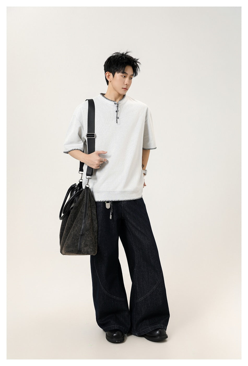 Model wearing a light gray short-sleeve henley with black contrast stitching and dark wide-leg jeans.