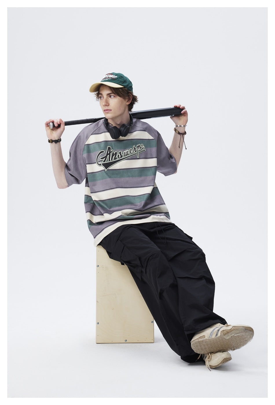 Seated model wearing a gray, teal, and cream striped oversized T-shirt with chest lettering and black cargo trousers.